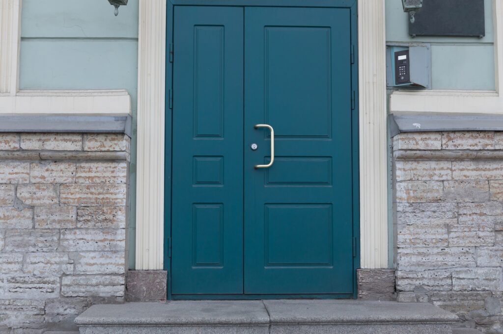 Blue double doors with a silver handle on a stone building entrance reflecting urban architecture. The walls are light-colored and feature decorative brickwork.