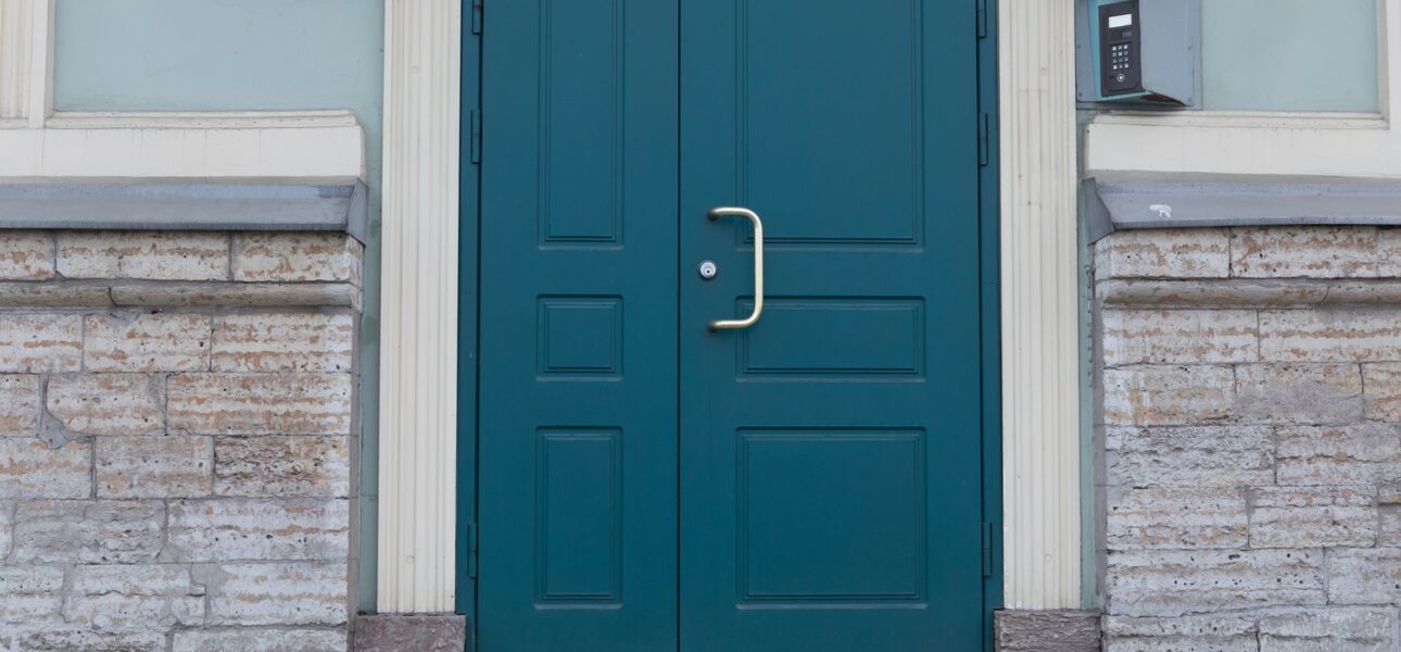 Blue double doors with a silver handle on a stone building entrance reflecting urban architecture. The walls are light-colored and feature decorative brickwork.